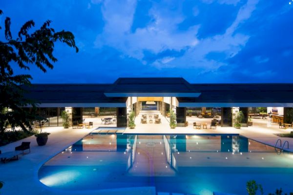 A large, luxurious swimming pool is illuminated in front of an elegant, modern building at twilight, surrounded by lounge chairs and greenery.