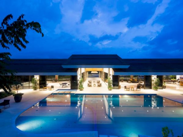 A large, luxurious swimming pool is illuminated in front of an elegant, modern building at twilight, surrounded by lounge chairs and greenery.