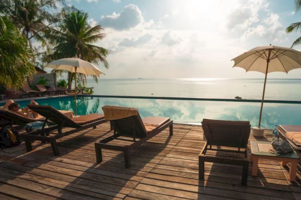 A serene poolside scene with lounge chairs, umbrellas, palm trees, and a view of the calm ocean under a partly cloudy sky at sunrise or sunset.