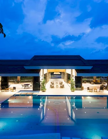 An illuminated modern building with an outdoor pool at dusk, featuring lounge areas and a large covered pavilion with open sides.