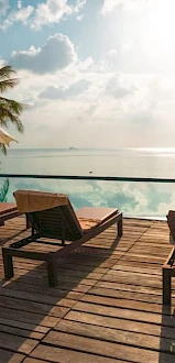 Sun loungers and umbrellas on a wooden deck by a tranquil pool, with palm trees and a serene ocean view under a partly cloudy sky.