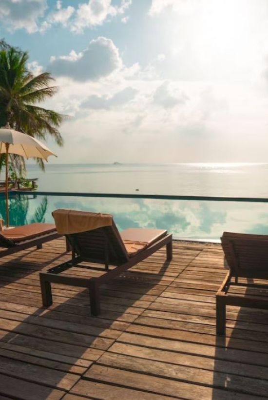 A serene poolside scene with lounge chairs and umbrellas on a wooden deck overlooking a calm body of water, framed by palm trees under a sunny sky.