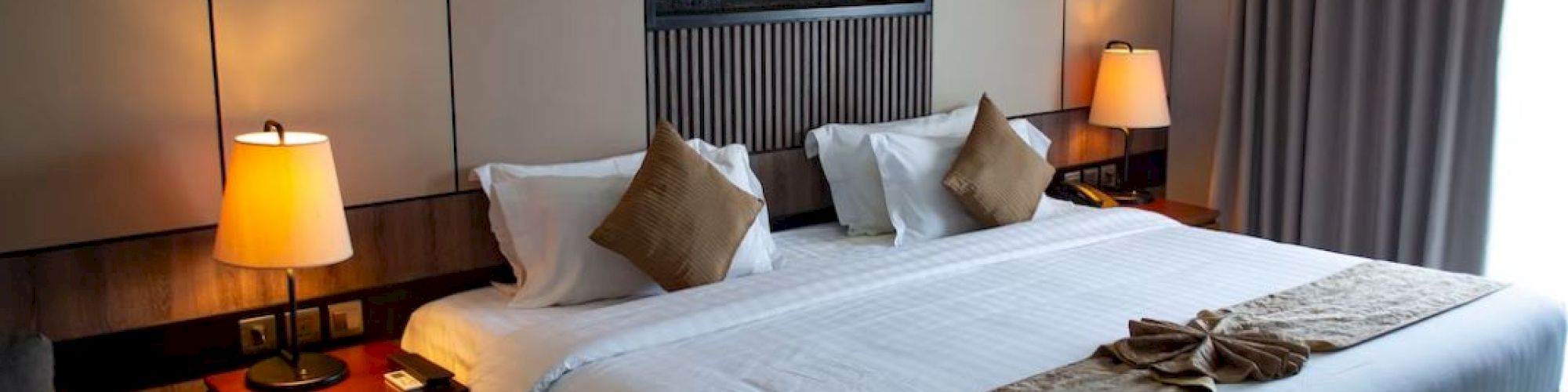 A modern hotel room with a large bed, bedside lamps, decorative pillows, a wall-mounted art piece, and floor-to-ceiling curtains.