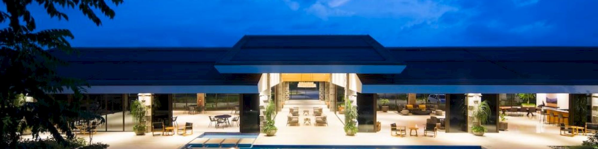 The image shows a spacious, illuminated outdoor pool area at dusk with a modern building in the background, featuring open seating and dining areas.