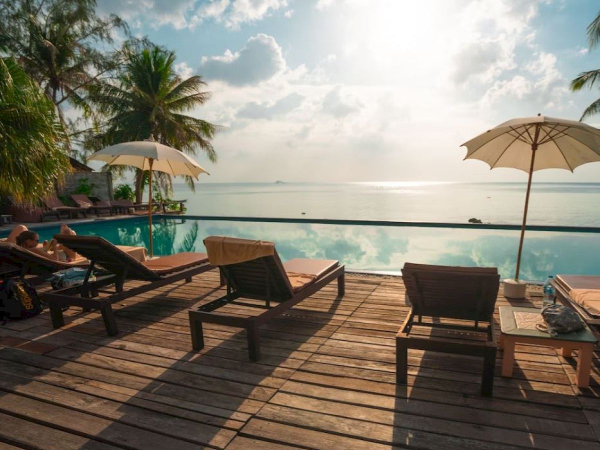 The image shows a tranquil poolside area with wooden lounge chairs, umbrellas, palm trees, and a view of the ocean under a partly cloudy sky.