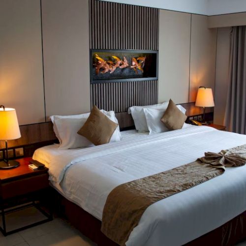 A neatly arranged hotel bedroom with warm lighting, featuring a large bed, pillows, nightstands with lamps, and curtains covering a window.