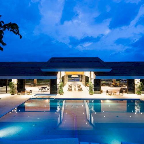 An illuminated outdoor swimming pool area in the foreground, with a modern building and open patio spaces in the background under a dusky sky.