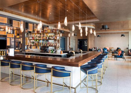 A modern bar scene with blue bar stools, a wooden countertop, an illuminated liquor shelf, and pendant lights, located in a spacious, stylish lounge.