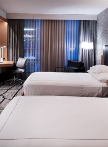 A modern hotel room features two neatly made beds, a wall-mounted TV, a desk with an office chair, and large windows with curtains.