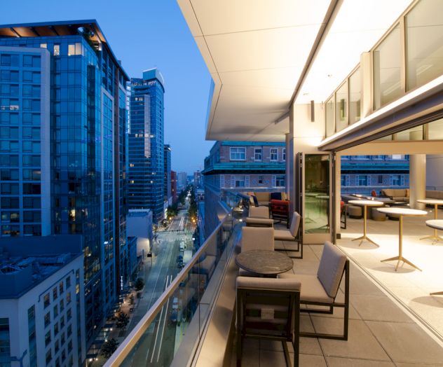 The image shows a modern urban balcony with seating and tables, overlooking a city street lined with tall buildings during twilight.