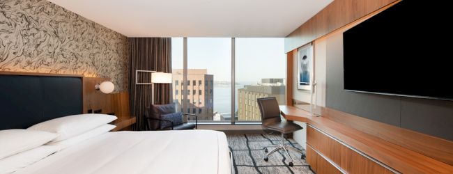A modern hotel room with a large bed, desk, chair, TV, and windows showcasing a city view is displayed in the image.