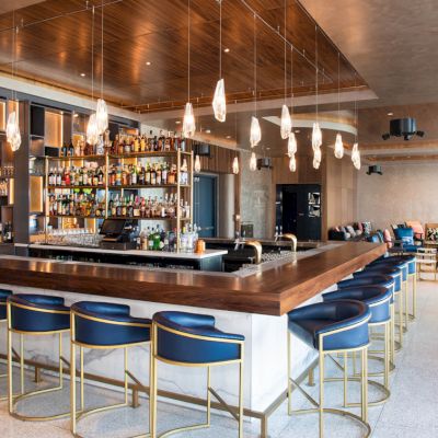 The image shows a modern bar with a wooden counter, blue chairs, hanging lights, and a fully stocked back bar, in a spacious and stylish interior.