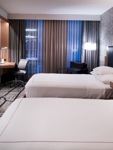 The image shows a modern hotel room with two neatly made beds, a wall-mounted TV, a work desk next to a window, and stylish decor.