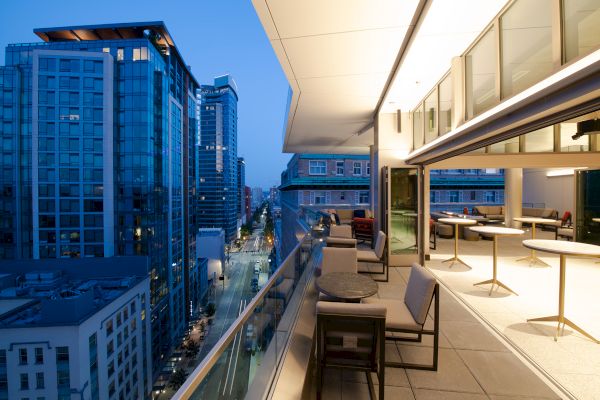 The image shows a cityscape view at dusk from a modern balcony with outdoor seating, overlooking tall buildings and a street below.