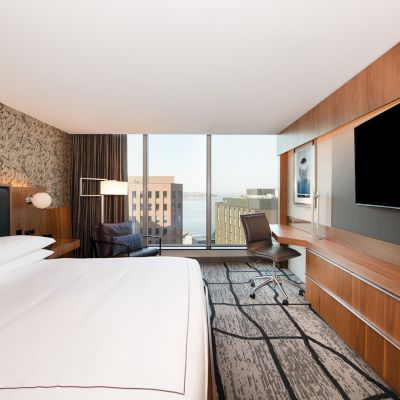 A modern hotel room with a large bed, wall-mounted TV, desk, chair, and a window view of city buildings and water in the background.