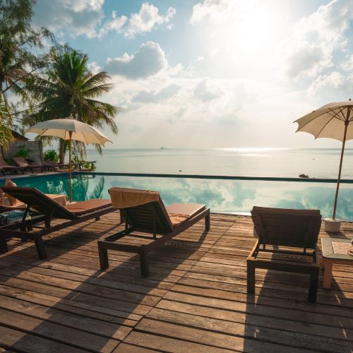 The image shows a tropical beach scene with lounge chairs and umbrellas on a wooden deck by a calm ocean at sunset.