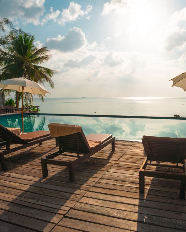 The image shows a tropical beach scene with lounge chairs and umbrellas on a wooden deck by a calm ocean at sunset.