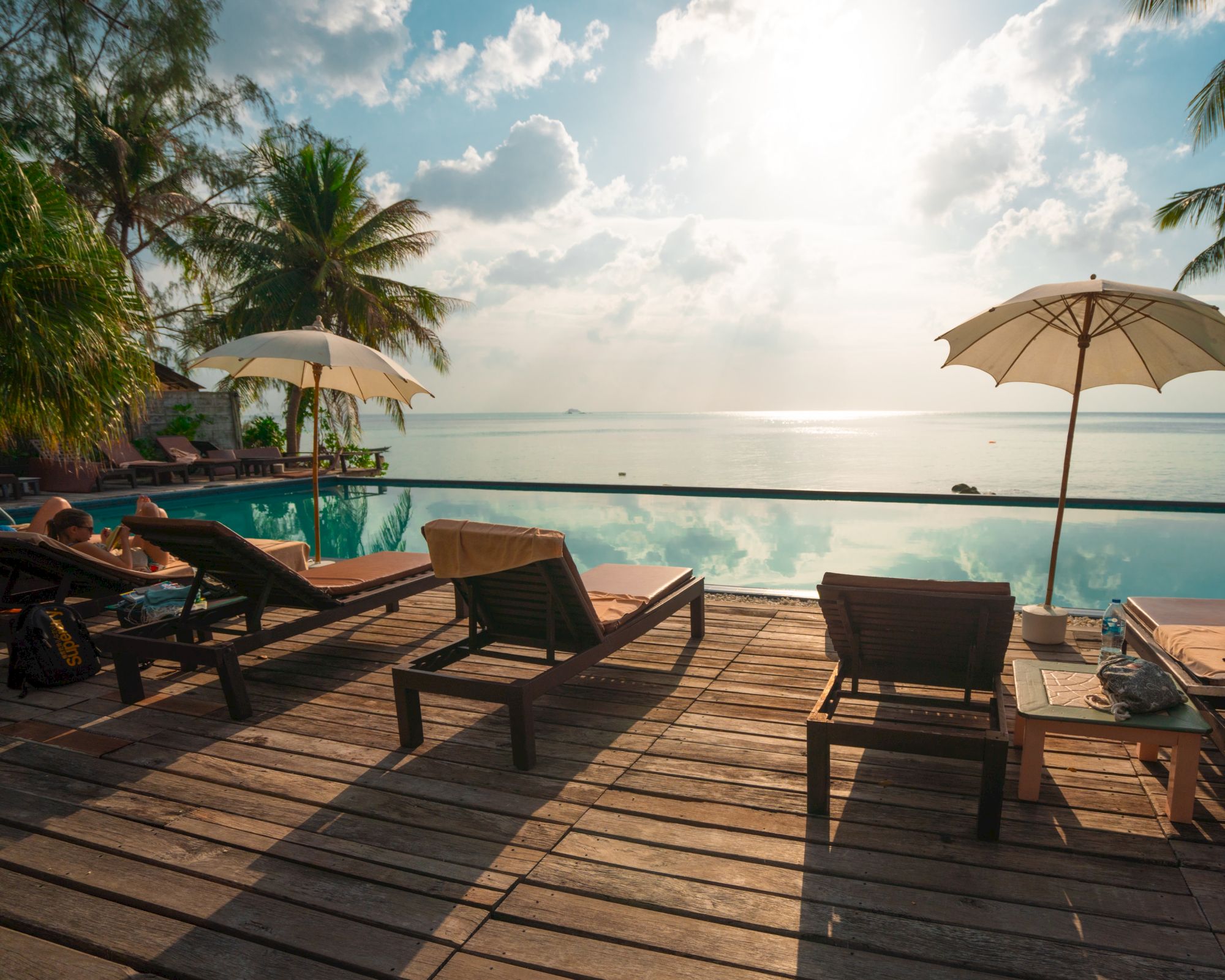 This image shows a tranquil beach scene with lounge chairs on a wooden deck, umbrellas, and a pool overlooking the serene ocean under a bright sky.