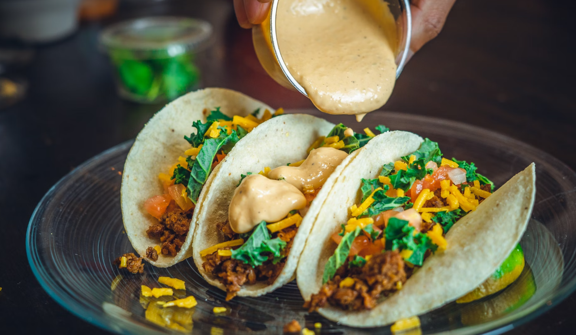 The image shows three tacos on a plate being drizzled with a creamy sauce. They are filled with ground meat, lettuce, tomatoes, and shredded cheese.