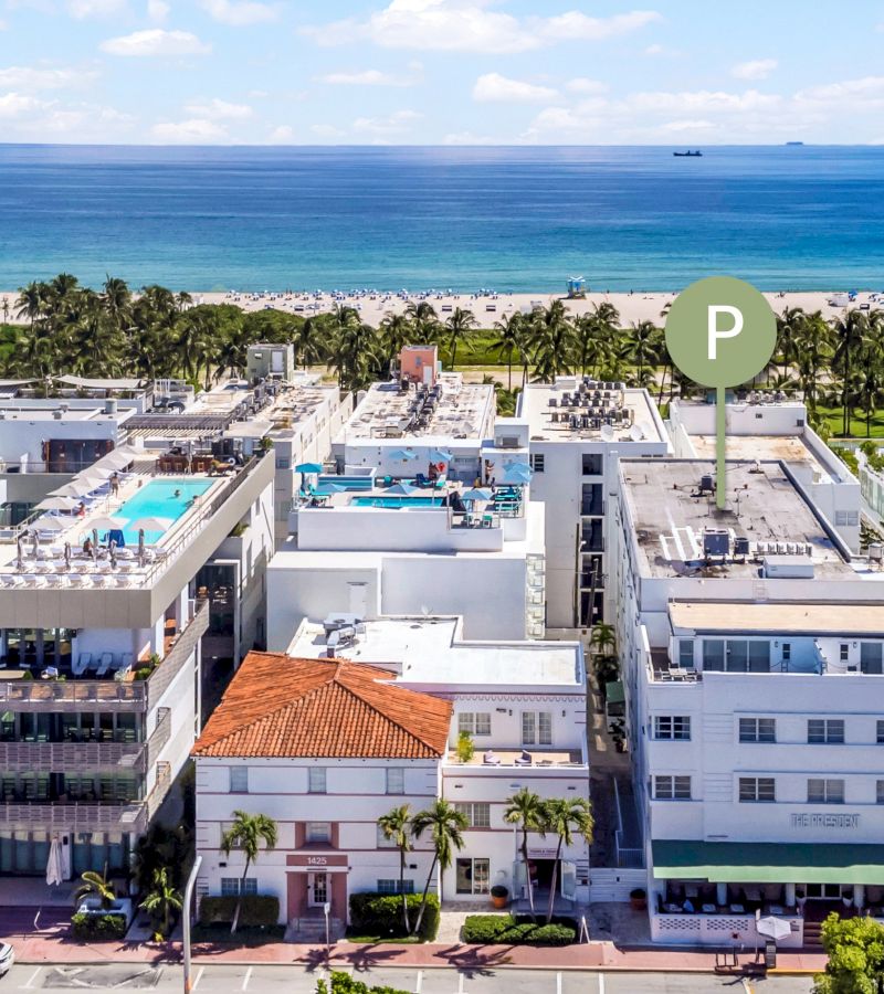 An aerial view of a white building with a green awning and a red-tiled roof, surrounded by palm trees and a parking lot, with a beach and ocean in the background.