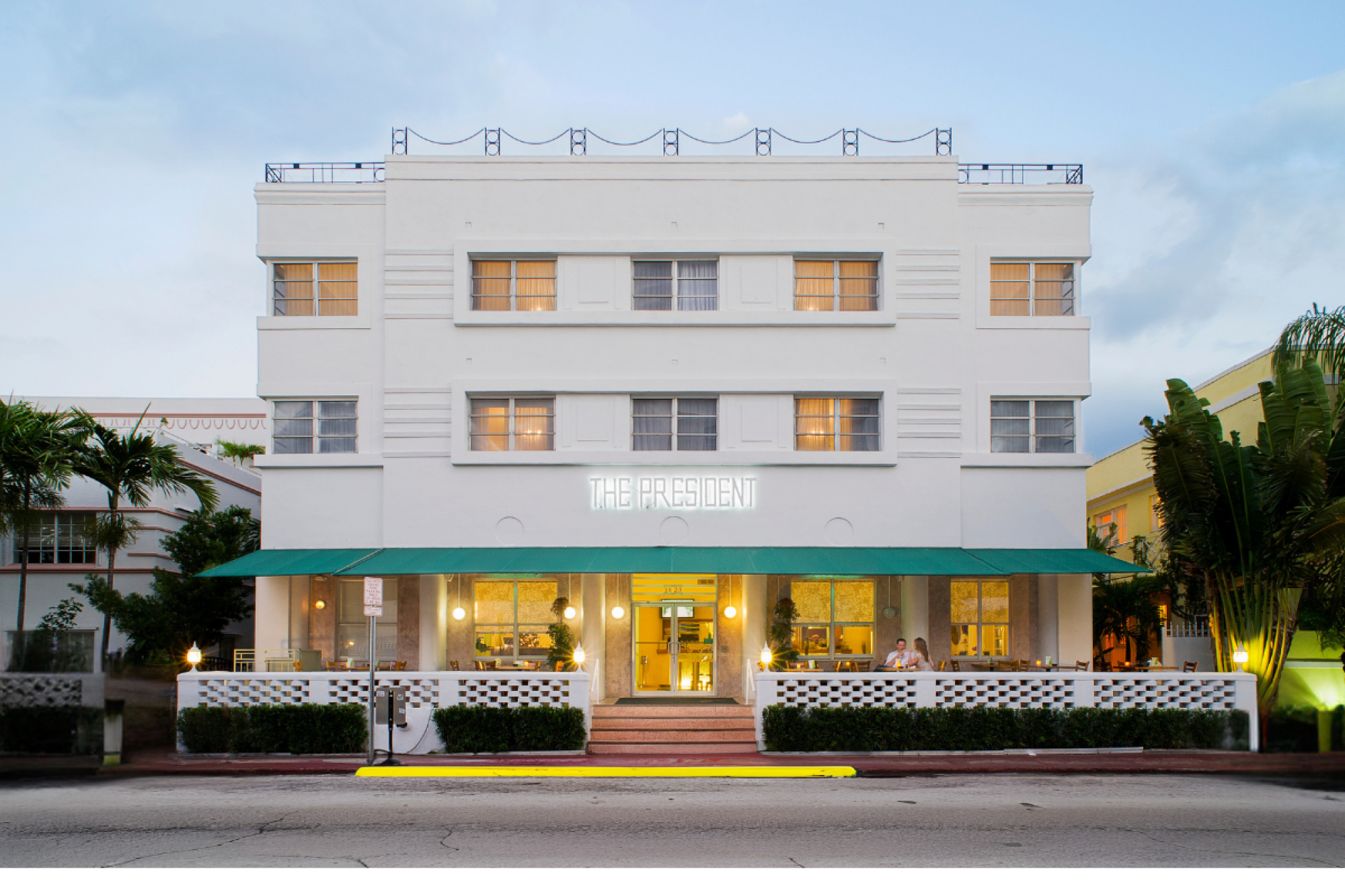 A three-story white building with a green awning over the entrance, surrounded by plants, seen from the street with a clear sky in the background.