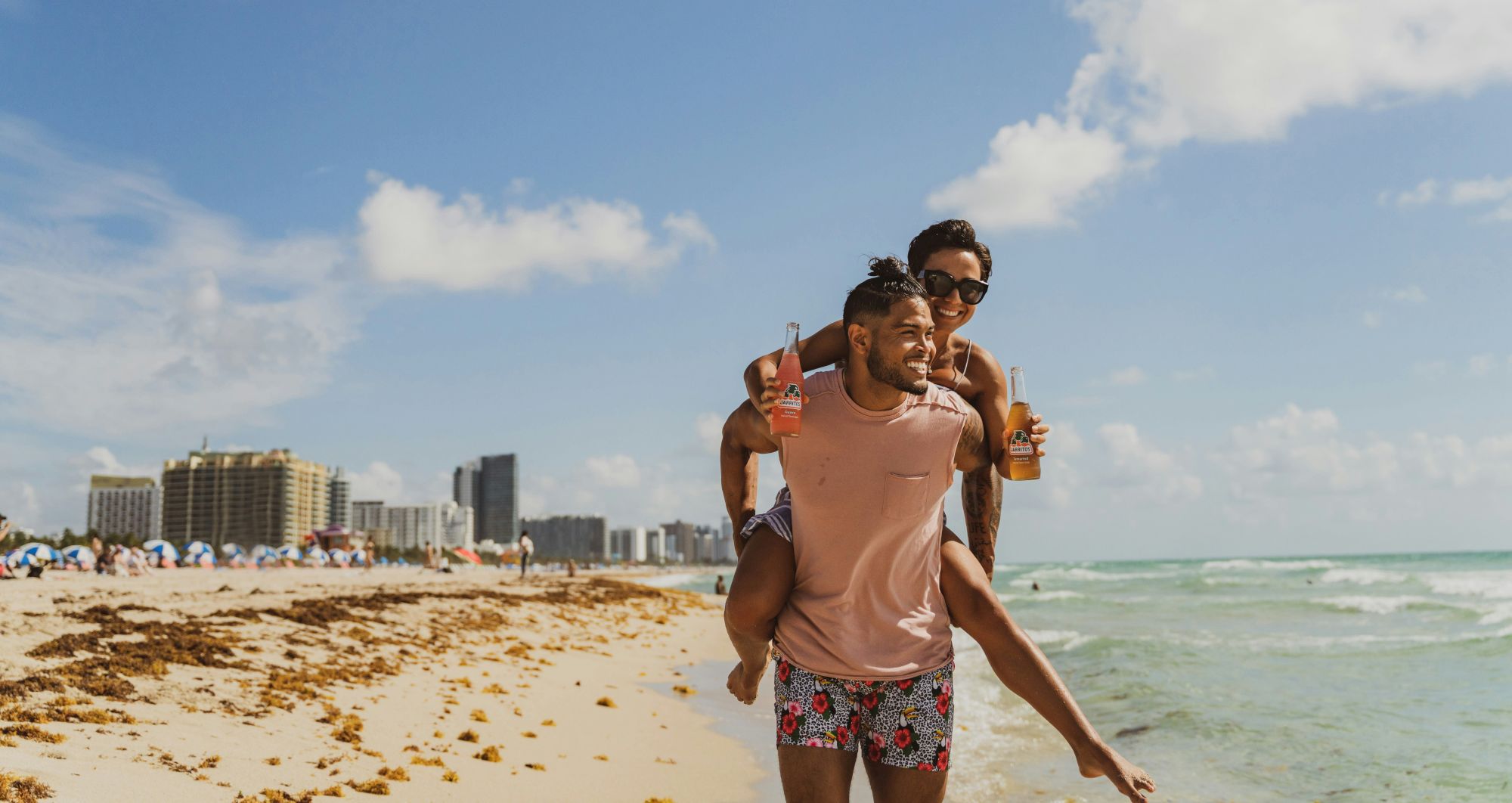 A couple enjoys a playful moment on a beach with one giving a piggyback ride, while holding drinks, with buildings and the ocean in the background.