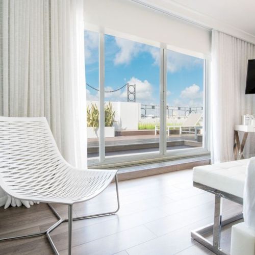 A modern, bright room with a white chair, bed, and TV. Floor-to-ceiling windows reveal a terrace and view of a bridge under a clear sky.