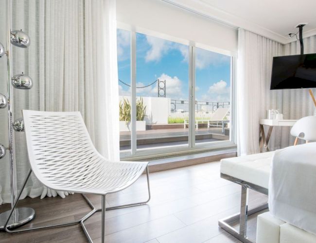 A modern, bright room with a white chair, bed, and TV. Floor-to-ceiling windows reveal a terrace and view of a bridge under a clear sky.