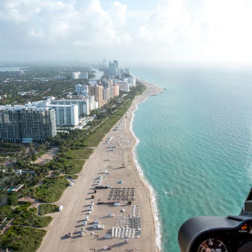 Aerial view of a beach with buildings on the left, umbrellas, and people on the sand, with a distant cityscape and turquoise water on the right.