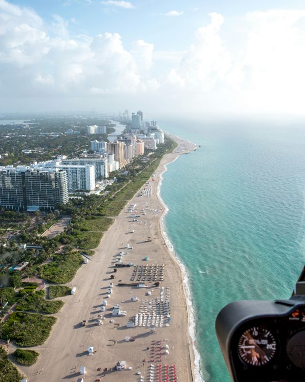 Aerial view of a beach with buildings on the left, umbrellas, and people on the sand, with a distant cityscape and turquoise water on the right.