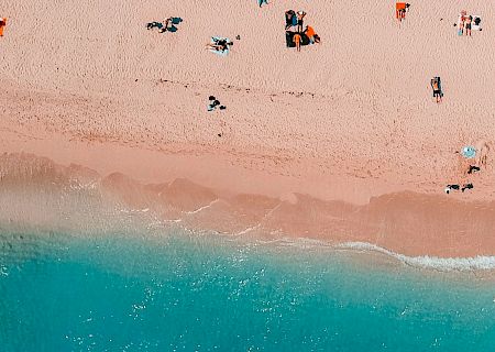 The image shows an aerial view of a beach with people relaxing on the sand near the turquoise ocean water.