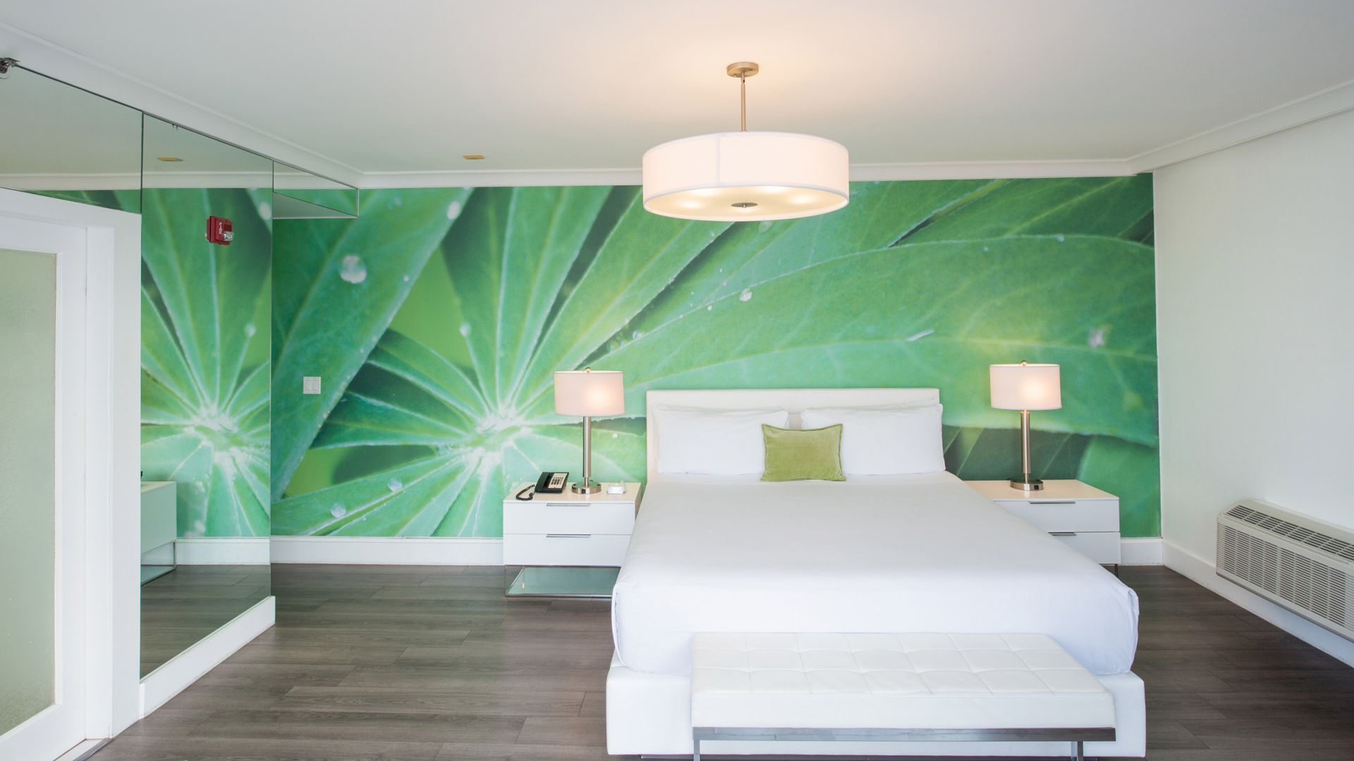 A modern bedroom with a large green leaf mural, white bed, and furniture, including two bedside lamps, a bench, and a mirrored wall on the left.