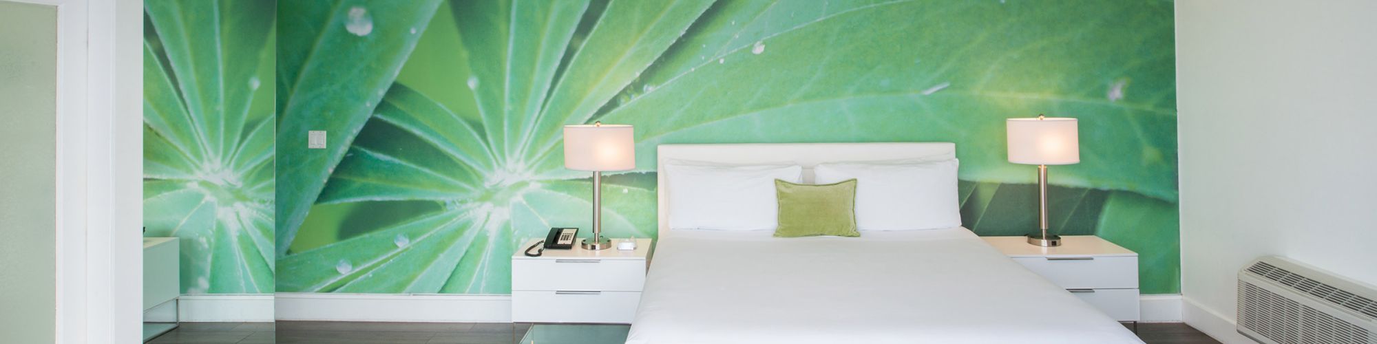 A modern bedroom with a large green leaf mural, white bed, and furniture, including two bedside lamps, a bench, and a mirrored wall on the left.