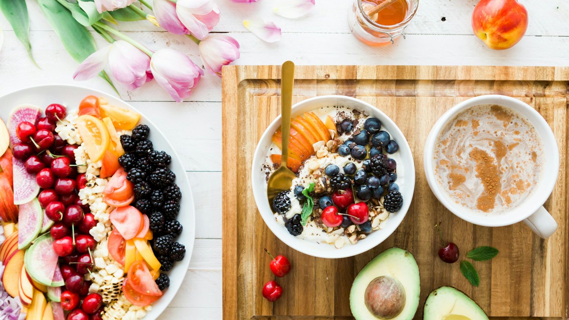 A breakfast spread with fruits, a bowl of granola, coffee, an avocado, and flowers on a white wooden table.