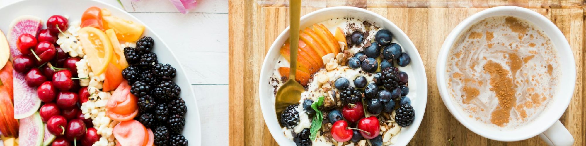 A breakfast spread with fruits, a bowl of granola, coffee, an avocado, and flowers on a white wooden table.