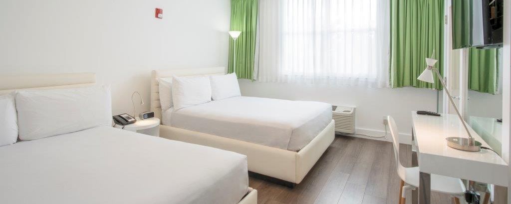 A hotel room with two beds, a desk, and a window with green curtains.