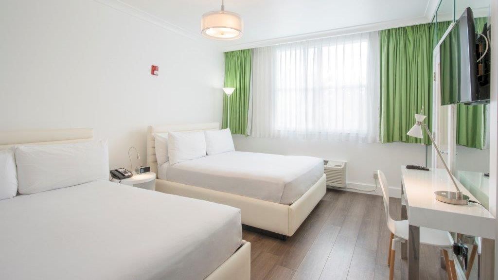 The image shows a hotel room with two beds, white bedding, green curtains, a TV on a wall, a desk, and a chair by the window.