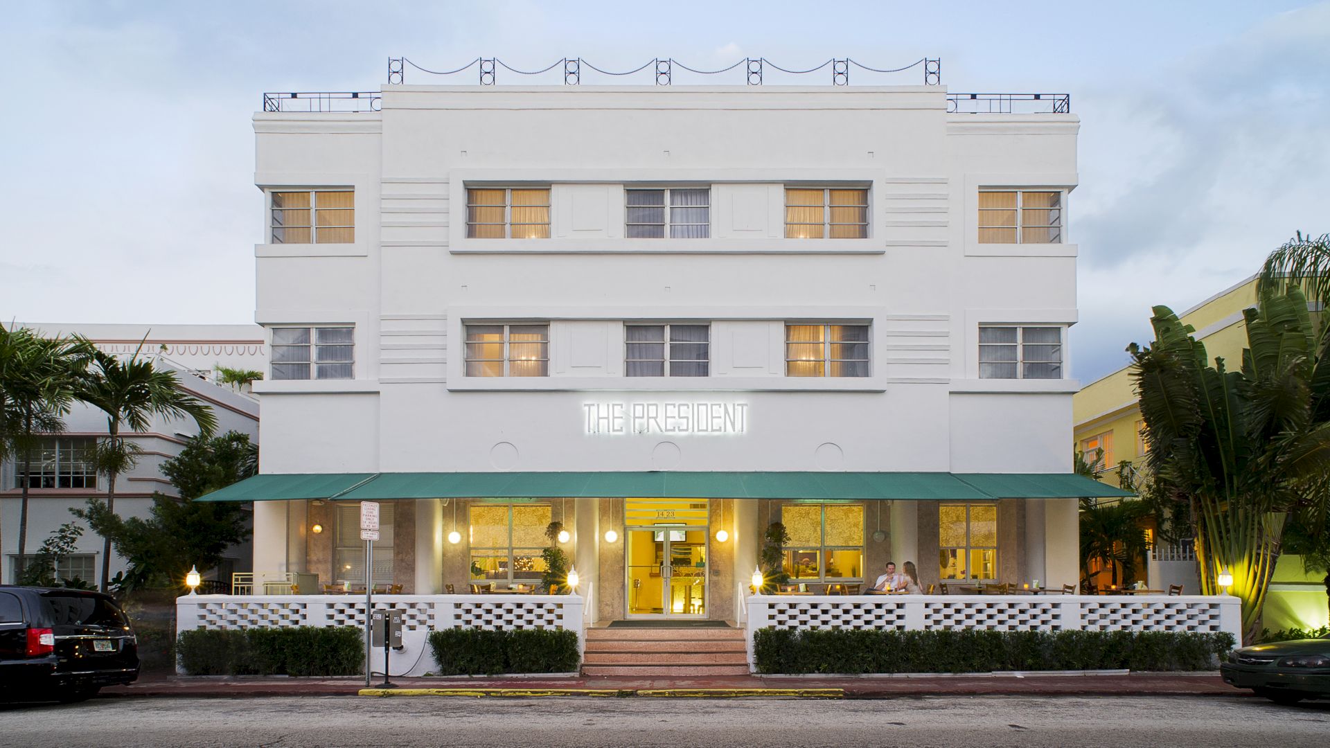 Front view of a white, three-story building named "THE PRESIDENT" with a green awning over the entrance and plants lining the front.
