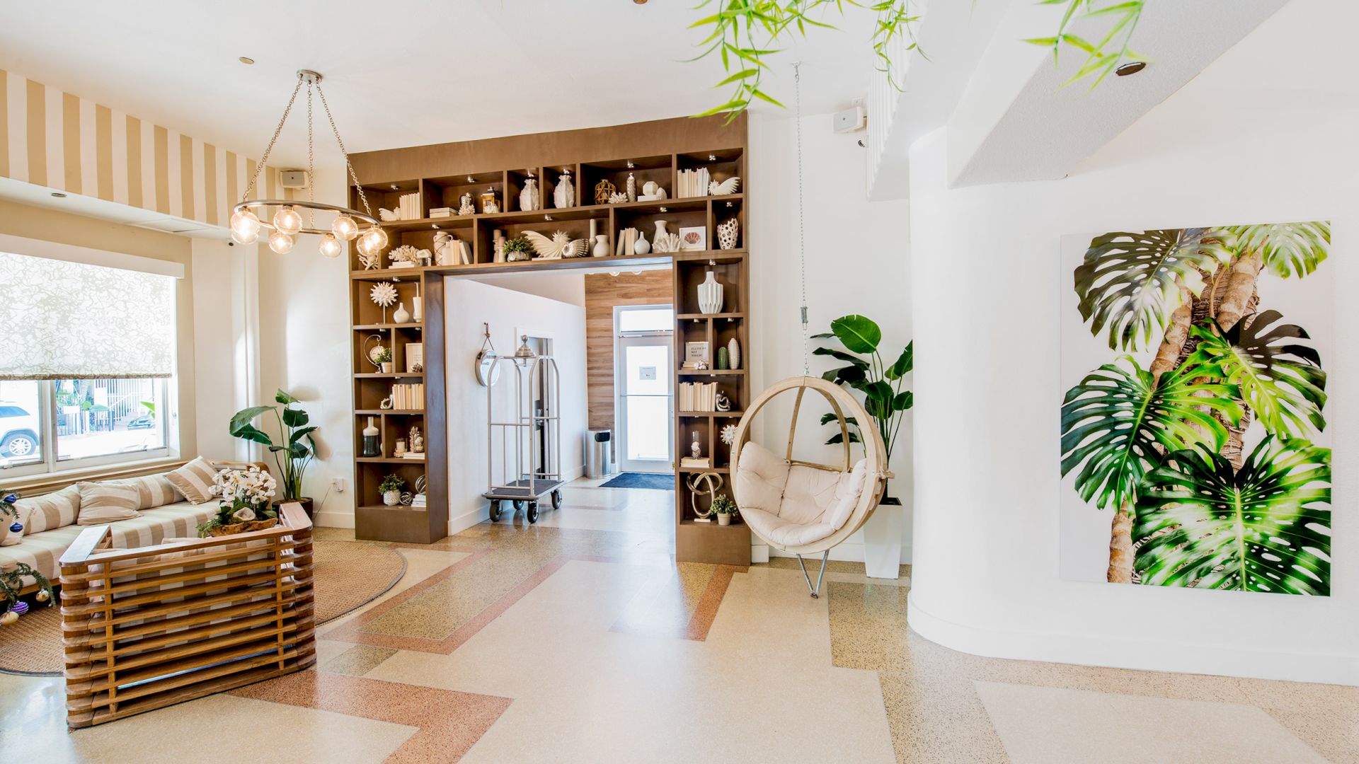 A stylish room featuring a cozy sitting area, hanging chair, bookshelf, modern chandelier, leafy decor, and large window with natural light.