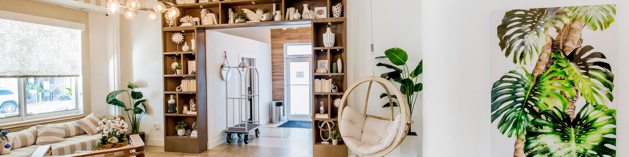 A stylish room featuring a cozy sitting area, hanging chair, bookshelf, modern chandelier, leafy decor, and large window with natural light.