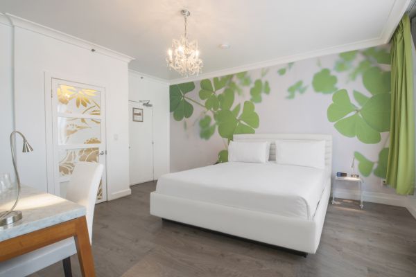 The image shows a modern bedroom with a large bed, white furniture, a chandelier, green leaf wall mural, and a decorative door featuring floral patterns.