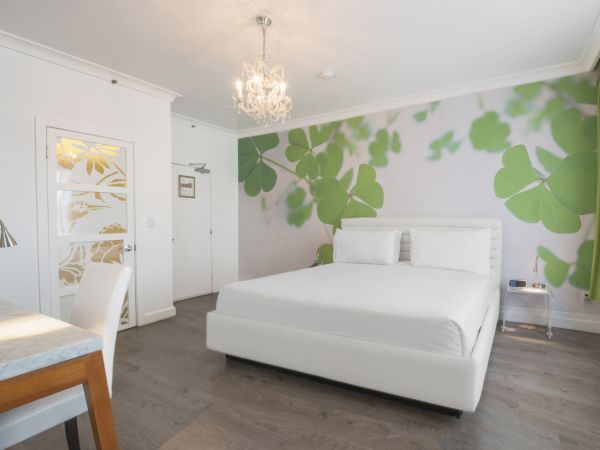 The image shows a modern bedroom with a large bed, white furniture, a chandelier, green leaf wall mural, and a decorative door featuring floral patterns.
