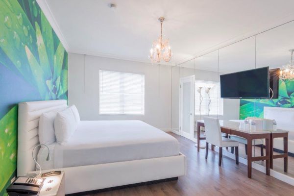 A modern, well-lit bedroom with a large bed, a green accent wall, a chandelier, a mirrored wall, a TV, a desk with chairs, and wooden flooring.