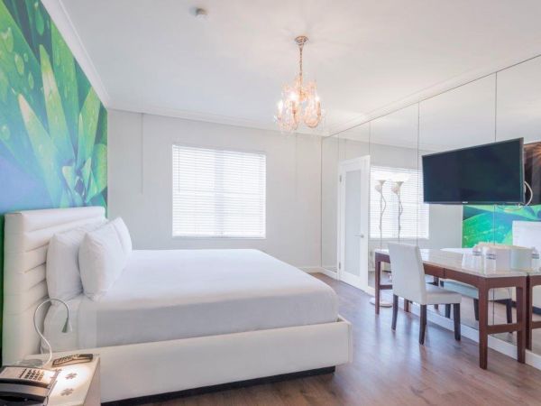 A modern, well-lit bedroom with a large bed, a green accent wall, a chandelier, a mirrored wall, a TV, a desk with chairs, and wooden flooring.