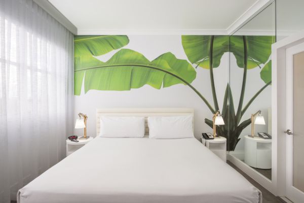 A modern bedroom with a large bed, tropical-themed wall mural, bedside tables with lamps, a mirrored wall, and a window with sheer curtains.