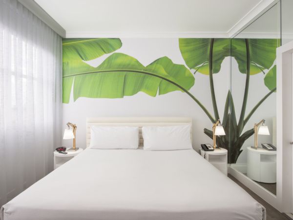 A modern bedroom with a large bed, tropical-themed wall mural, bedside tables with lamps, a mirrored wall, and a window with sheer curtains.