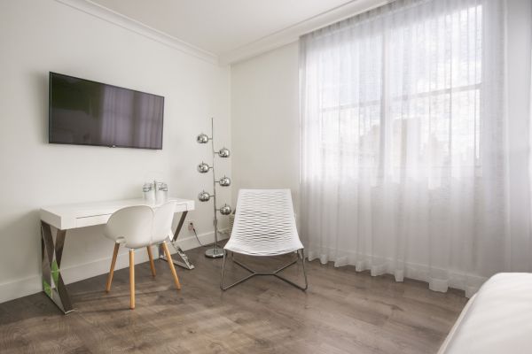 A modern room with a white desk, chair, TV, lamp, window with sheer curtains, and a minimalist chair with wooden flooring, creating a clean look.