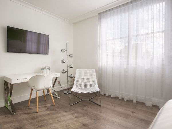 A modern room with a white desk, chair, TV, lamp, window with sheer curtains, and a minimalist chair with wooden flooring, creating a clean look.