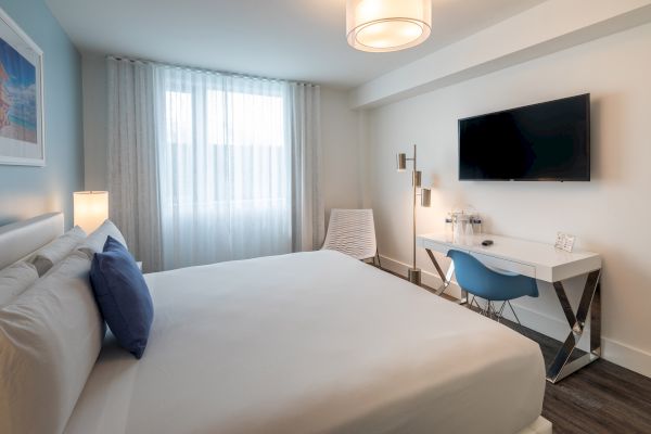 A modern hotel room with a large bed, blue-accented decor, a desk with a blue chair, a wall-mounted TV, and soft lighting filtering through sheer curtains.