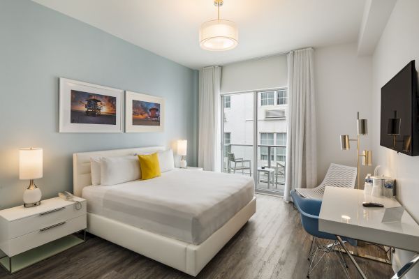 This image shows a modern hotel room with a bed, desk, TV, and two nightstands. The room features minimalistic decor and a large window.